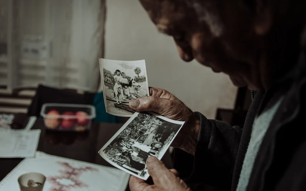 Um senhor sentado a uma mesa, examinando fotografias antigas em preto e branco sob
a luz de uma janela.
