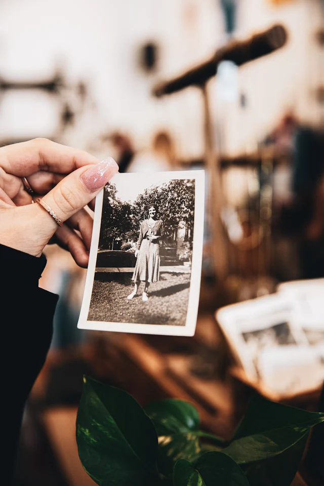 Uma mão segurando uma fotografia antiga em preto e branco de uma mulher, com o
              restante do álbum desfocado ao fundo.